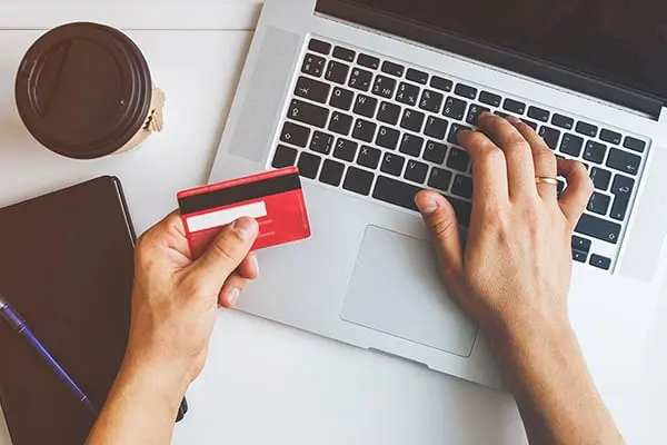 Overhead view of a person's hands as they hold a red credit card in one hand and navigate on a laptop with the other, possibly making an online purchase or completing a payment.